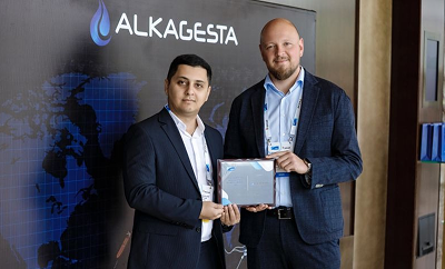 Two individuals standing indoors in front of an Alkagesta-branded backdrop while holding a document together.