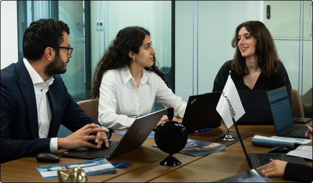A group of office team members seated around a meeting table with laptops and documents, collaborating in a modern workspace.