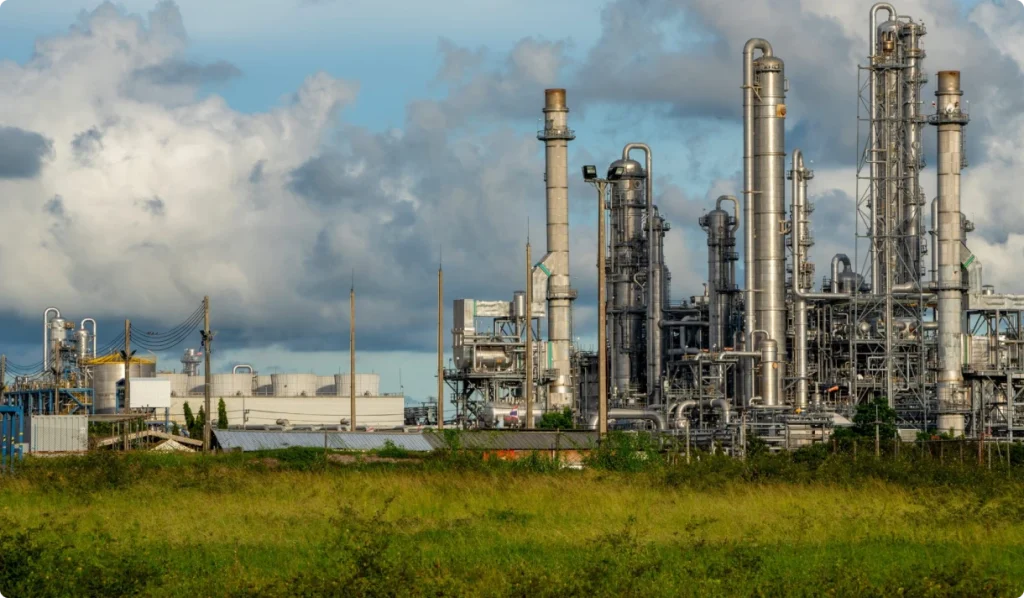 An industrial refinery complex with multiple tall metal towers, pipes, and processing structures, set against a cloudy sky with green grass in the foreground.