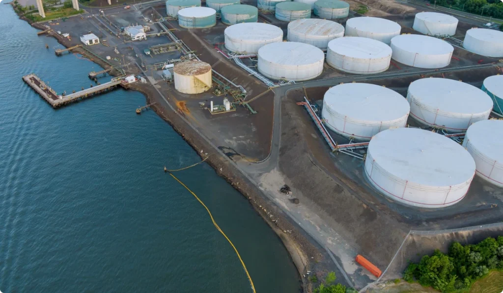 An aerial view of a coastal industrial storage facility with multiple large white storage tanks arranged across the site near the shoreline.
