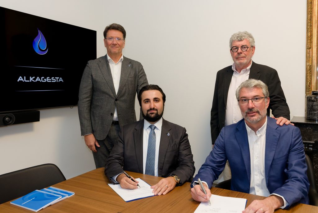 Four individuals in business attire seated and standing around a table during a formal document signing session in a corporate meeting room with an Alkagesta logo displayed on a screen.