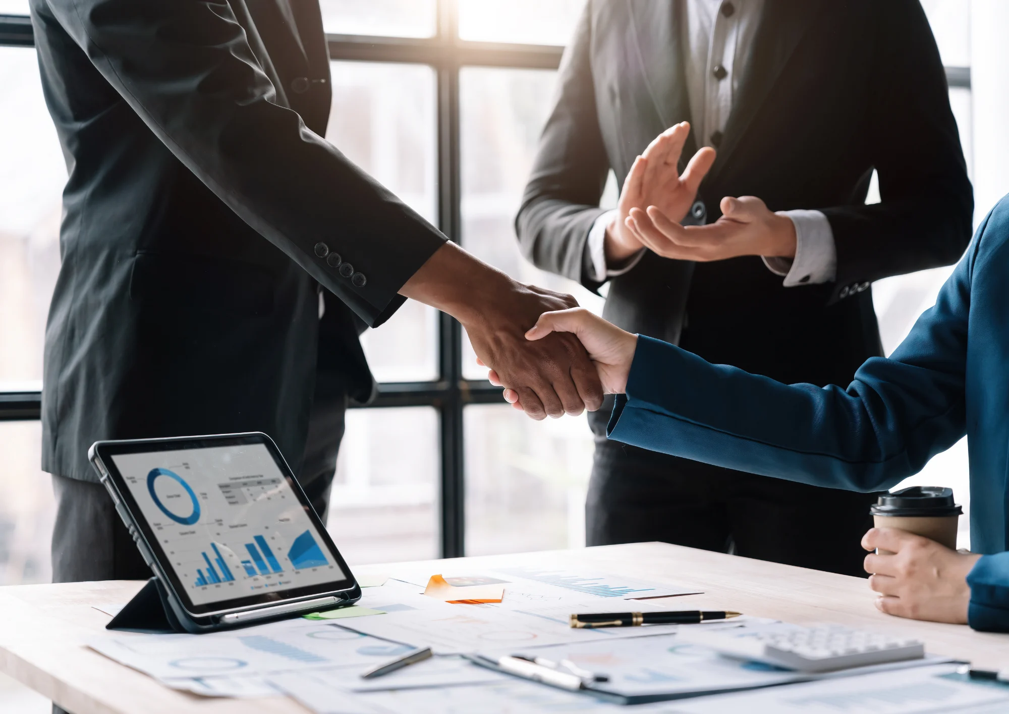 Two people shaking hands over a meeting table with documents, charts, and a laptop.