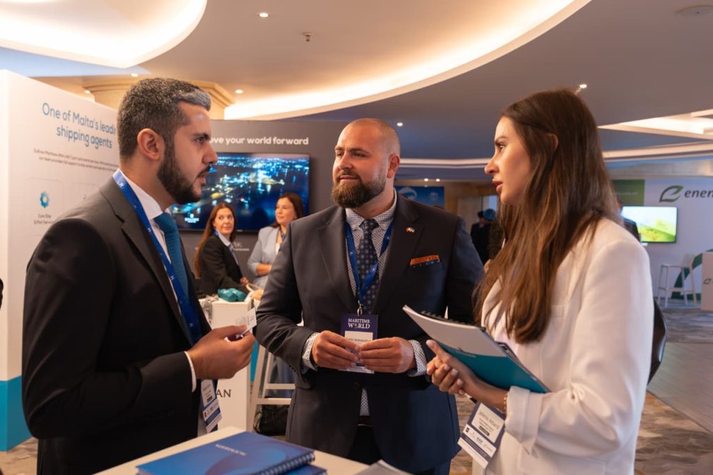 Three individuals standing in a conference exhibition area having a discussion, with event booths, banners, and displays visible in the background.