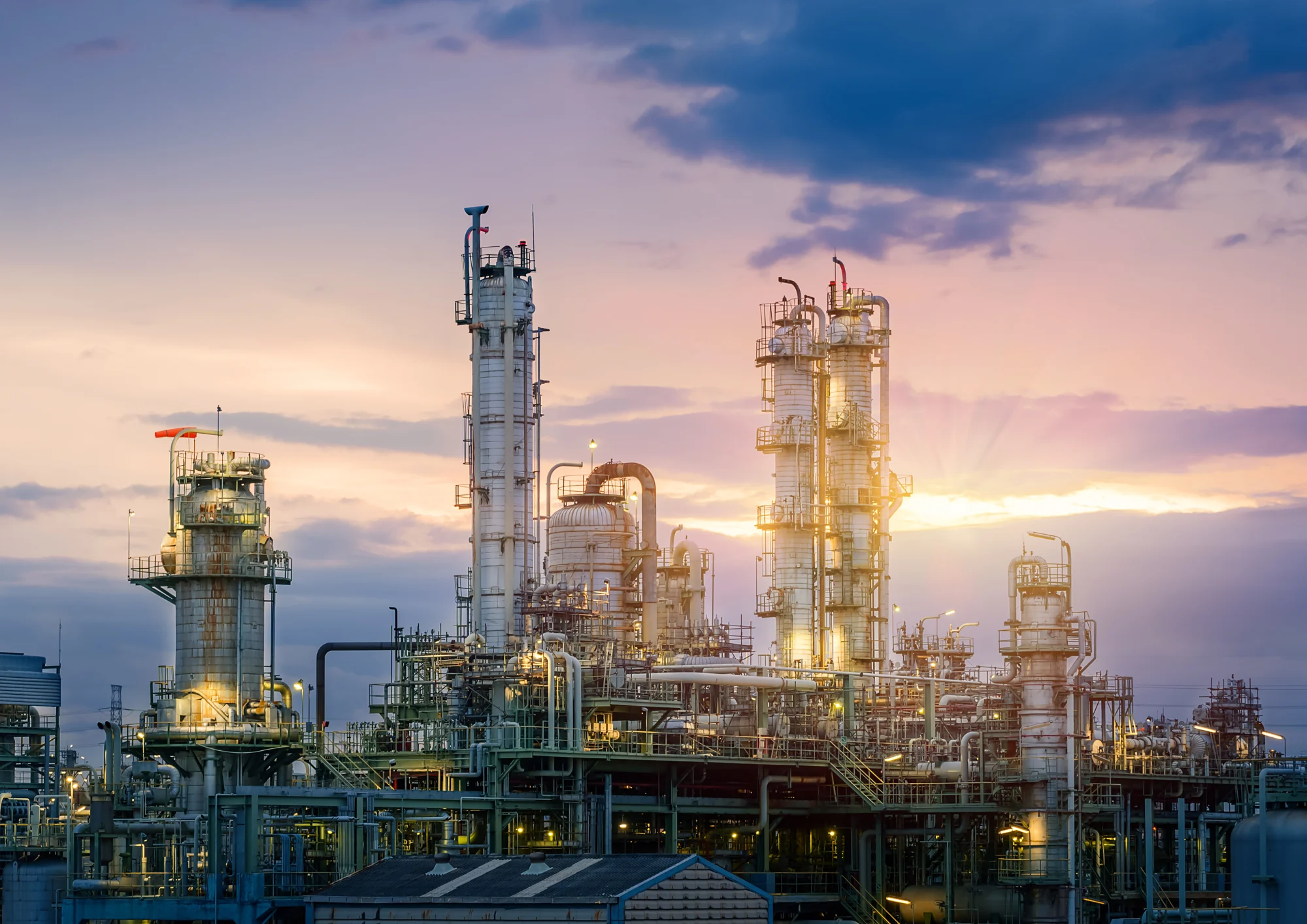 An industrial refinery with multiple towers, pipes, and metal structures illuminated at sunset.