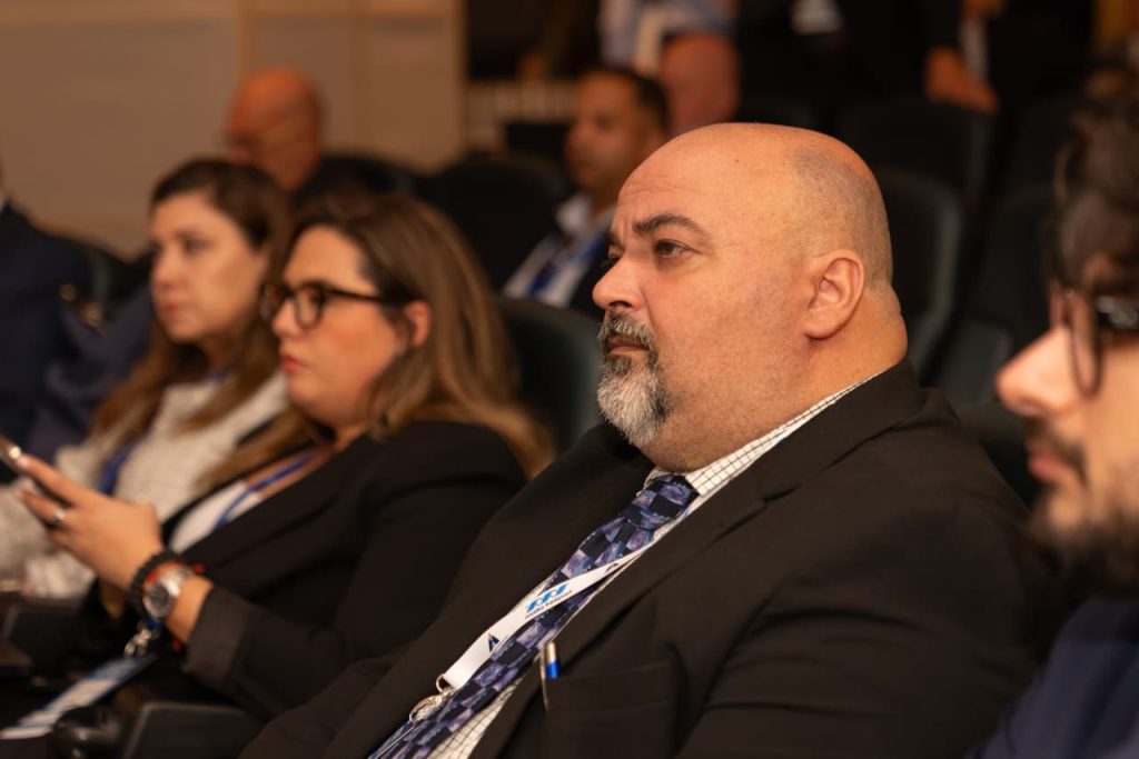 A group of attendees seated in rows during a conference session, facing forward with notepads and lanyards visible.