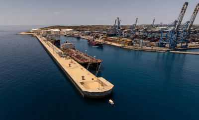 An aerial view of a long pier extending into the water with a large vessel docked alongside, and cranes and port infrastructure visible in the background.