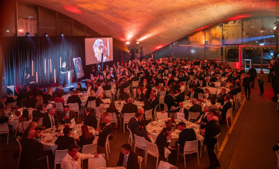 A large formal event with many attendees seated at round tables inside a spacious venue with warm overhead lighting and a stage area visible at the front.