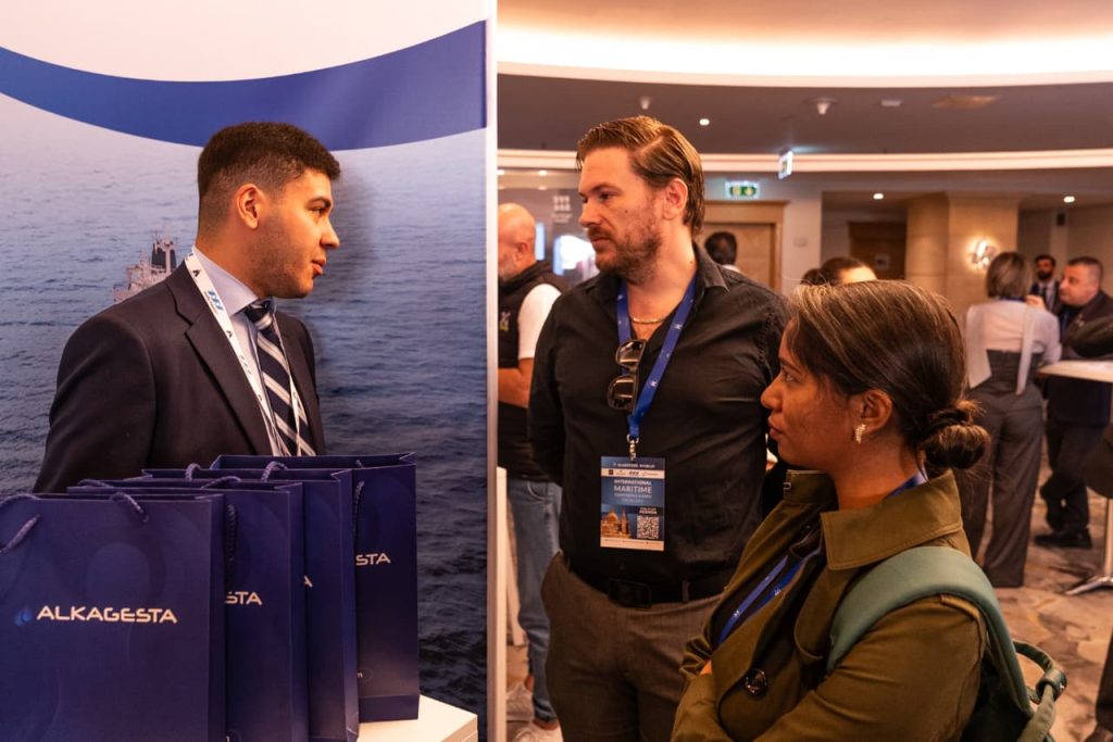A person standing at an Alkagesta exhibition booth beside branded bags, speaking with two event attendees in a conference hall.