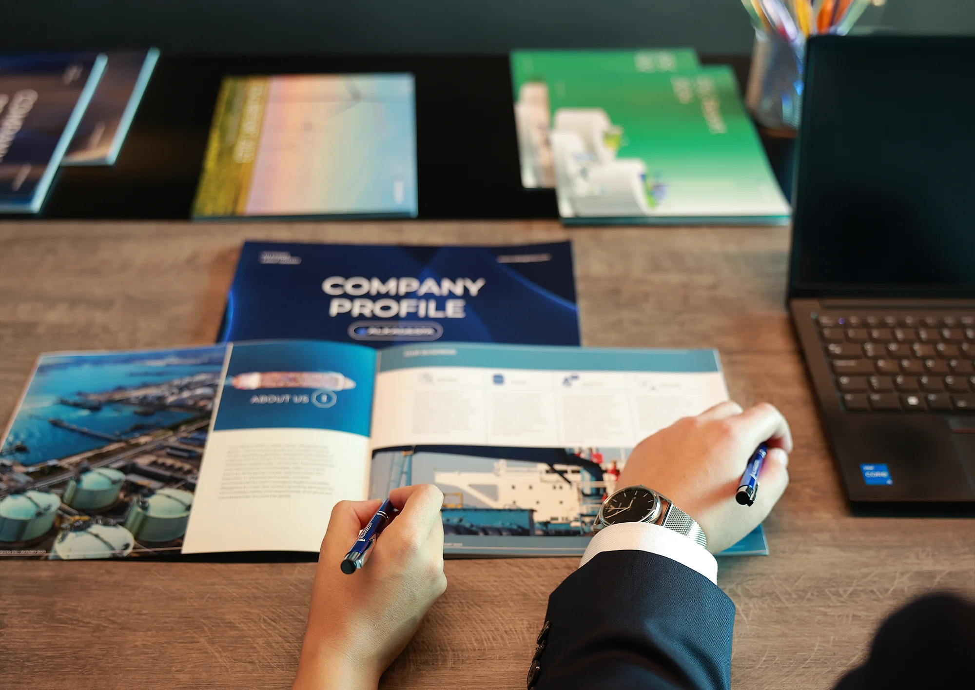A person reviewing a company profile booklet on a desk alongside documents, brochures, and a laptop.