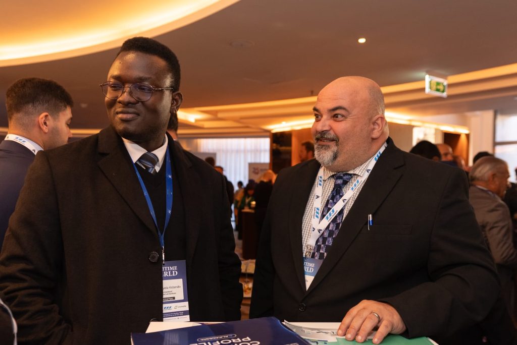 Two individuals standing at an Alkagesta event area, wearing conference badges, with branded banners and displays in the background.