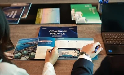 People sitting around a meeting table reviewing a company profile booklet and other printed materials, with a laptop and documents arranged on the desk.