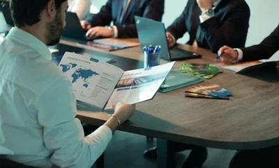 People seated around a meeting table reviewing printed charts and documents, with laptops and notebooks placed on the table.