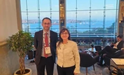 Two individuals standing together in an indoor event area with large windows overlooking a city and water view.
