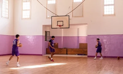 A basketball practice session inside a gym where three individuals are moving across the court under natural light from large windows.