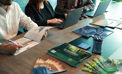 A meeting table with individuals seated around it, reviewing printed brochures, documents, and laptops.