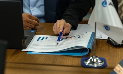 A close‑up of a hand pointing at charts inside an open booklet on a wooden meeting table, with a laptop and an Alkagesta flag nearby.