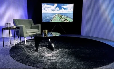 A studio setup with two chairs, a small table, and a large screen displaying an image of a ship, arranged on a circular rug with vertical panel lighting behind.
