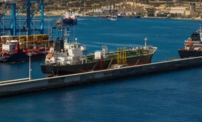 A large industrial port with multiple cargo ships docked alongside cranes and containers, with coastal buildings visible in the background.