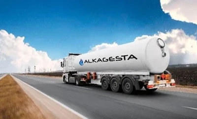 A large Alkagesta-branded tanker truck driving on a highway under a bright blue sky with clouds.