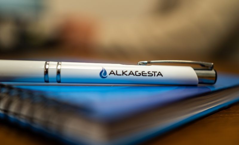 A close-up of an Alkagesta-branded pen resting on top of a closed blue notebook placed on a wooden desk.