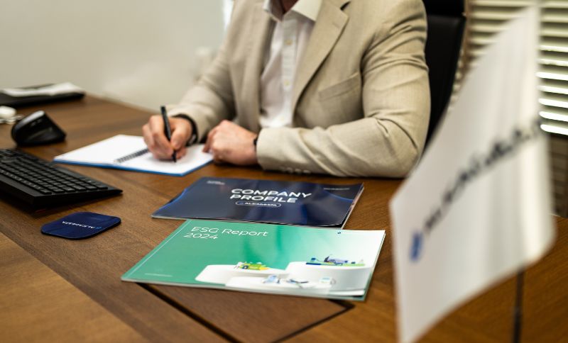 A person seated at a wooden desk writing on a document, with company brochures, a keyboard, and an Alkagesta-branded desk flag visible.