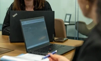 A meeting setting with individuals seated at a table using laptops and writing on documents, with notebooks and office supplies visible on the desk.