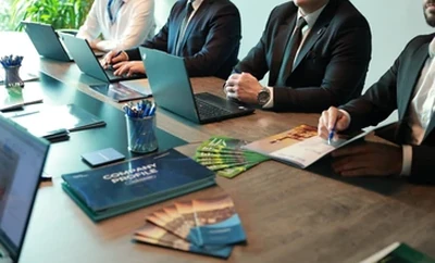 A group of individuals seated around a meeting table with laptops, documents, notebooks, and pens visible, reviewing materials in a professional office setting.