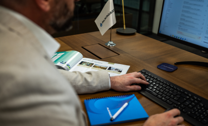A person seated at a wooden desk using a computer keyboard with documents, a blue notebook, a pen, and a small Alkagesta flag placed nearby.