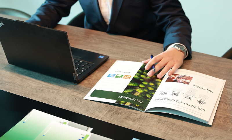 A person seated at a desk with an open environmental brochure, a laptop, and additional documents spread out across the wooden surface.