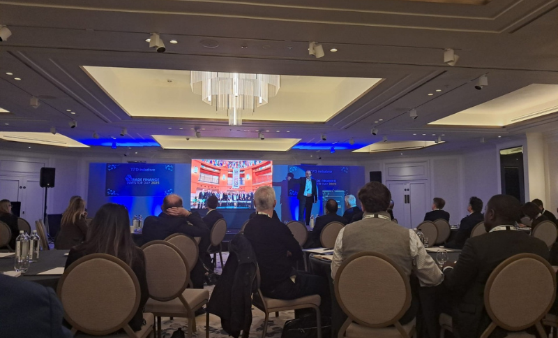 A conference room with attendees seated at round tables facing a stage where a presentation is being shown on large screens.