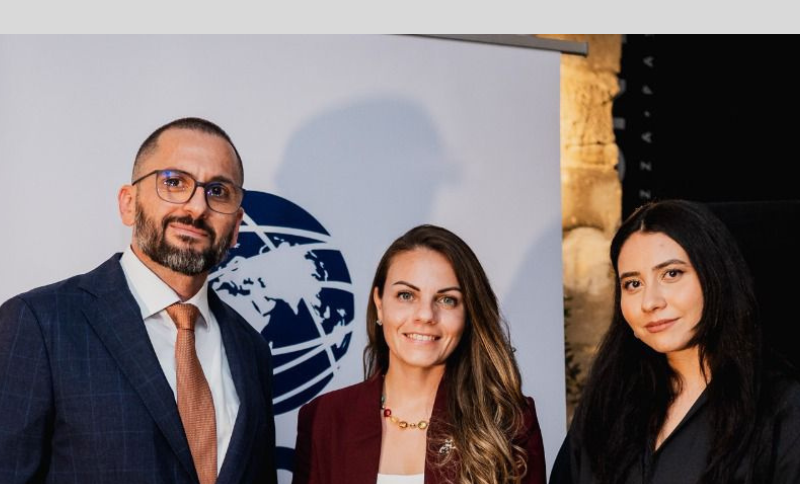 Three individuals standing in front of a backdrop with a circular maritime-themed logo, posing together in an event setting.