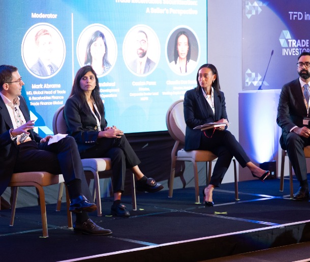A conference panel with several speakers seated on a stage in front of a large screen displaying moderator and speaker information.