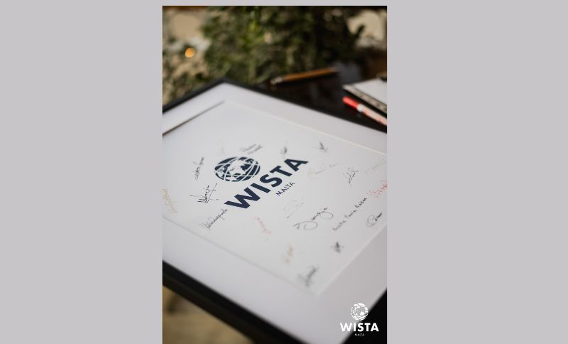 A framed WISTA document placed on a table, featuring printed signatures around the WISTA logo, with pens and greenery visible in the background.