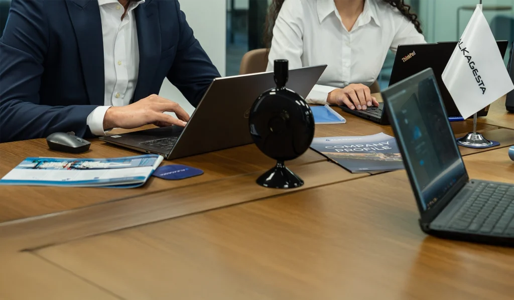 A meeting table with open laptops, a conference microphone, printed brochures, and an Alkagesta tabletop flag, with several individuals seated around the table.