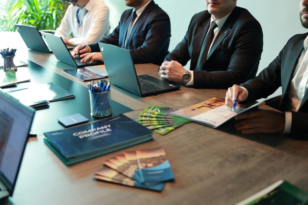 A meeting table with individuals seated around laptops, printed compliance documents, brochures, notebooks, and pens during an office work session.