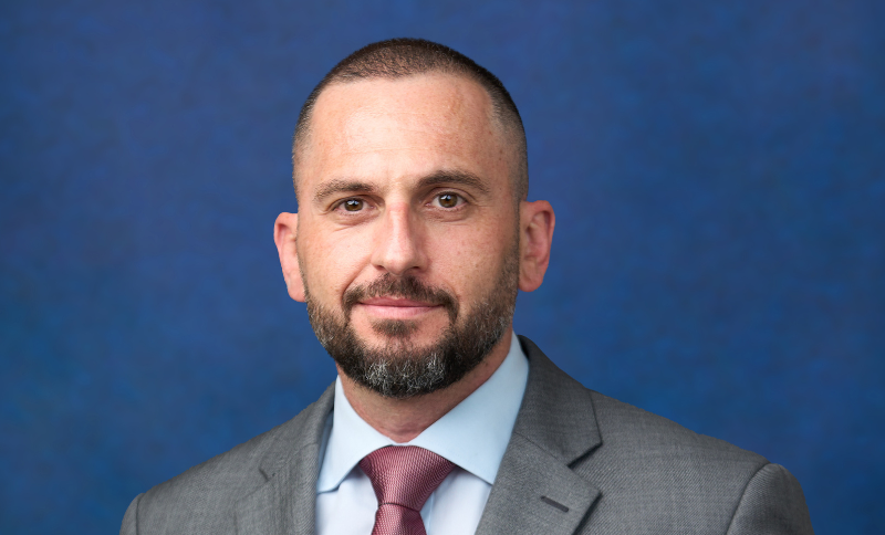 A portrait of an individual wearing a suit, positioned in front of a solid blue background.