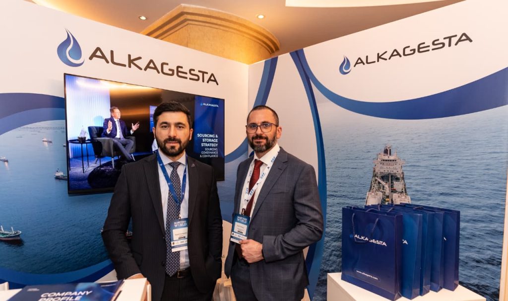 Two individuals standing at an Alkagesta exhibition booth with branded banners, a display screen, and promotional bags visible in the background.