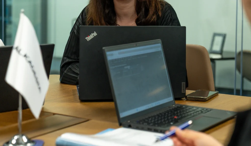 A meeting table with laptops, documents, notebooks, and an Alkagesta-branded flag, with individuals seated and working in an office setting.