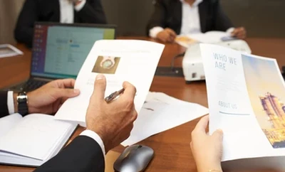 Hands holding and reviewing printed documents at a meeting table, including a booklet with charts and an image on the cover.