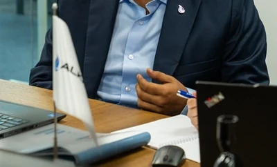 A meeting table with an individual seated nearby, holding a pen and reviewing documents next to a laptop, an Alkagesta-branded desk flag, and other office materials.