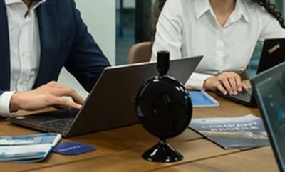 A meeting table with individuals using laptops and reviewing printed documents, with an Alkagesta-branded desk microphone placed at the center.