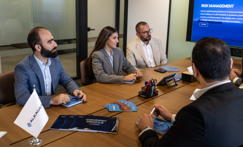 A group of people sitting around a meeting table with documents, notebooks, and a screen displaying a risk management slide.