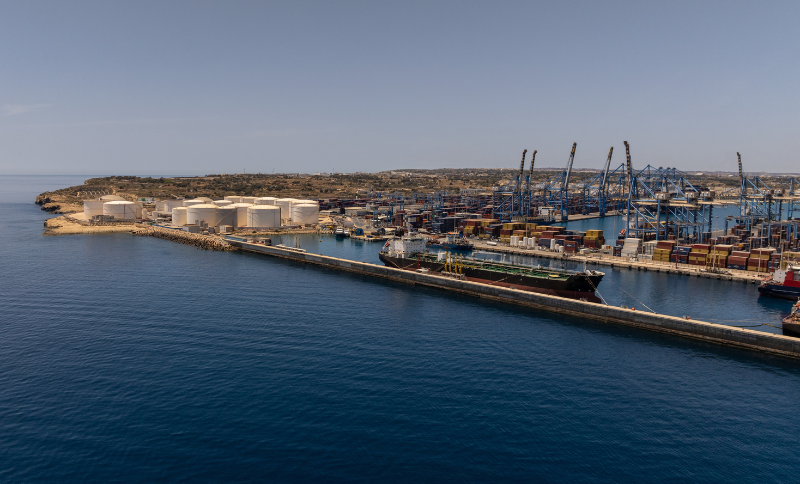 An aerial view of a large industrial port with storage tanks, cranes, and docked ships along the waterfront.