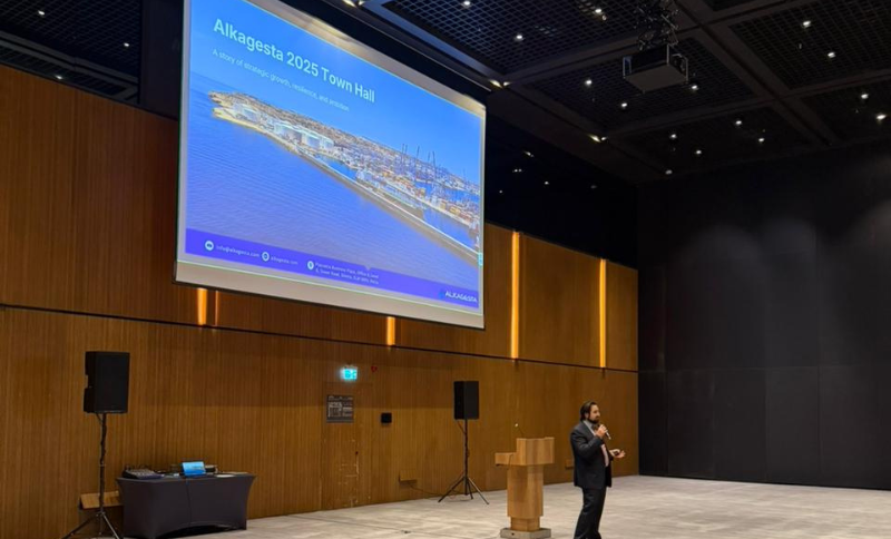 Alkagesta leader presenting on stage at 2025 Town Hall at Pervenire with large screen displaying Alkagesta 2025 Town Hall opening slide and maritime port image