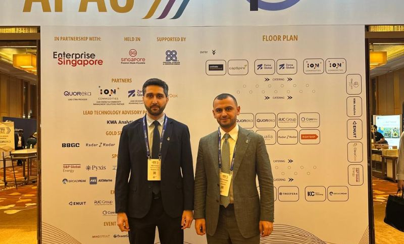 Two individuals in business attire standing in front of a Commodity Trading Week APAC event backdrop featuring sponsor logos and a floor plan.