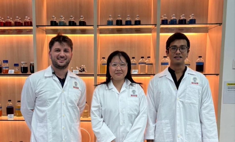 Three individuals wearing laboratory coats standing in front of illuminated shelves containing sample bottles inside a laboratory facility.