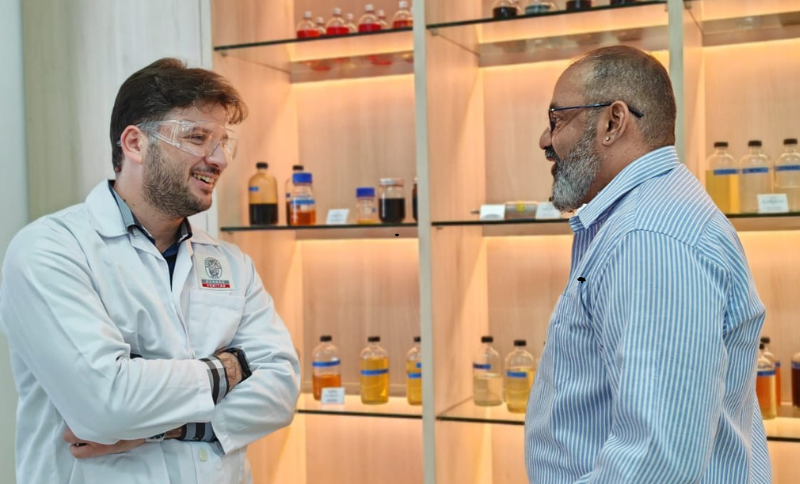 Two individuals wearing laboratory coats and protective eyewear standing in front of illuminated shelves holding bottles with various liquid samples inside a laboratory facility.