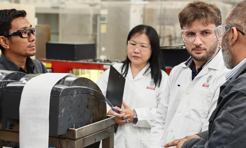 Team of individuals in safety goggles and laboratory coats observing industrial testing equipment inside an inspection facility.