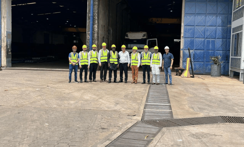 A group of eleven people wearing safety vests and helmets stand in a line outside a large industrial building with blue doors.