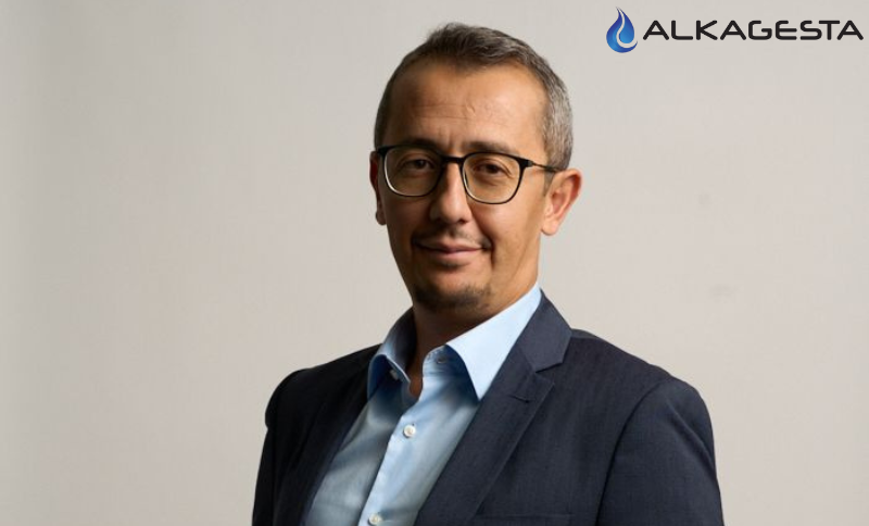 Individual in business attire standing in front of an Alkagesta-branded backdrop during a professional photoshoot.